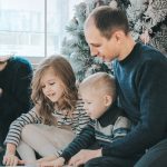 girl and boy reading book sitting between man and woman beside Christmas tree